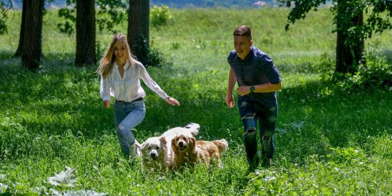 A joyful couple running with their dogs through a lush green field on a sunny day.